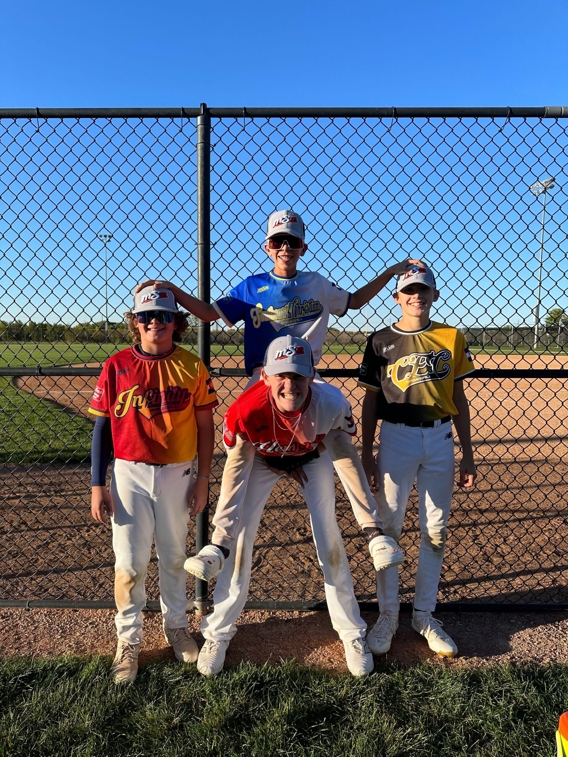 Four baseball players in different school-color jerseys pose beside a ballfield fence
